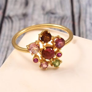 Könnte beinhalten: Ein Goldring mit einer Ansammlung verschiedenfarbiger Edelsteine in unterschiedlichen Größen. Die Steine in den Farben Dunkelrot, Pink, Hellgrün und Braun sind kreisförmig auf einer dünnen, polierten Goldschiene mit glatter Oberfläche angeordnet.