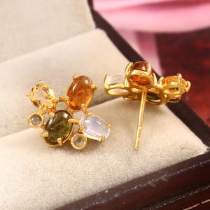 Könnte beinhalten: Ein Paar goldene Ohrstecker mit einer Ansammlung von mehrfarbigen ovalen und runden Edelsteinen, darunter gelbe, orangefarbene, olivgrüne und durchscheinende weiße Steine, gefasst in einen goldfarbenen Metallrahmen mit einem Stiftverschluss.