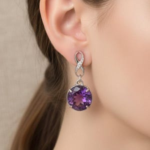 Könnte beinhalten: Nahaufnahme eines silbernen Ohrrings mit einem großen, runden, lila Edelstein. Der Ohrring hat ein dekoratives Unendlichkeitszeichen-Design an der Oberseite, das mit kleinen, klaren Steinen verziert ist. Der Edelstein hat eine tiefe, satte lila Farbe.