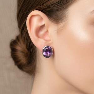 Könnte beinhalten: Ovale Amethyst-Ohrstecker mit Silberfassung. Die Ohrringe zeigen einen leuchtend violetten Edelstein. Ein klassisches Schmuckstück.