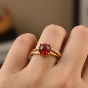 Könnte beinhalten: Ein goldener Ring mit einem großen, runden, roten Edelstein als Mittelpunkt. Das Band teilt sich in zwei Teile, wobei eine Seite mit kleinen, klaren Edelsteinen besetzt ist. Der Ring befindet sich an einem Finger.