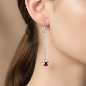 Könnte beinhalten: Silberner Ohrhänger mit einem zentralen roten Edelstein, umgeben von kleinen weißen Steinen in einem floralen Design. Der Ohrring hat eine lange, zarte Kette mit kleinen, funkelnden Akzenten.