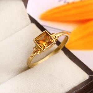 Könnte beinhalten: Ein goldener Ring mit einem quadratischen, bernsteinfarbenen Edelstein. Der Ring hat ein schlichtes Band mit dekorativen Details in der Nähe der Edelsteinfassung. Der Ring wird in einer weißen Schmuckschatulle mit verschwommenem orangefarbenem Hintergrund präsentiert.