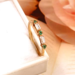 Könnte beinhalten: Ein goldener Ring mit Smaragd- und Baguette-Diamant-Akzenten. Der Ring hat ein zartes Band und ist mit drei runden Smaragden und zwei rechteckigen Diamanten besetzt. Die Edelsteine sind in einer Reihe auf der Oberseite des Rings angeordnet.