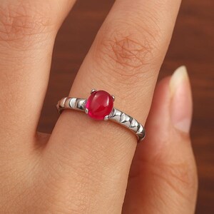 Könnte beinhalten: Ein silberfarbener Ring mit einem mittig platzierten, ovalen, glatten und leuchtend roten Edelstein. Die Ringschiene weist ein markantes, strukturiertes Muster aus einer Reihe sich wiederholender, leicht erhabener, halbkreisförmiger Metallsegmente auf.