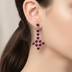 Könnte beinhalten: Hängeohrringe mit silberfarbenem Metallrahmen und leuchtend roten Edelsteinen. Die Ohrringe haben ein lineares Design mit einem kreuzförmigen Boden und zeigen einen klassischen und eleganten Stil.