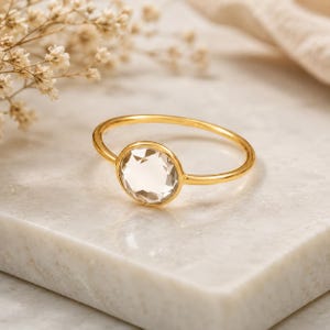 Könnte beinhalten: Ein goldener Ring mit einem einzelnen, runden, facettierten, klaren Edelstein in einer einfachen Zargenfassung. Das Band ist dünn und poliert und zeigt ein schlichtes Design, das die Klarheit und das Funkeln des zentralen Steins hervorhebt.