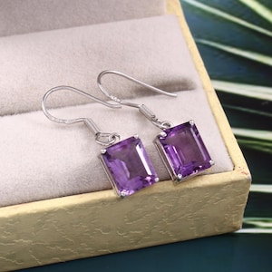 Könnte beinhalten: Ein Paar silberner Ohrringe mit rechteckigen, tiefvioletten Amethyststeinen. Die Ohrringe werden in einer hellbeigen Schmuckschatulle präsentiert. Die Haken haben ein einfaches, gebogenes Design.