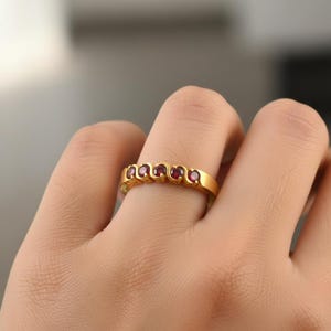 Könnte beinhalten: Ein goldfarbener Ring mit fünf kleinen, runden, roten Edelsteinen, die in einem wellenförmigen Muster angeordnet sind. Der Ring befindet sich an einem Finger, der Hintergrund ist unscharf.