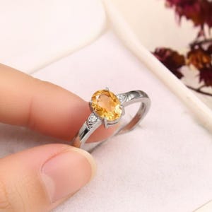 Könnte beinhalten: Ein silberner Ring mit einem ovalen, gelben Edelstein als Mittelpunkt. Der Ring hat zwei kleine, klare Edelsteine auf beiden Seiten des Hauptsteins. Der Ring ist vor einem weißen Hintergrund platziert.