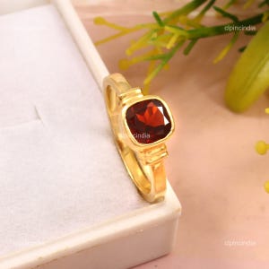 Könnte beinhalten: Ein goldener Ring mit einem quadratischen, tiefroten Edelstein. Der Ring hat ein schlichtes Band und eine quadratische Fassung. Der Ring wird auf einer weißen Oberfläche mit grünem und gelbem Laub im Hintergrund präsentiert.