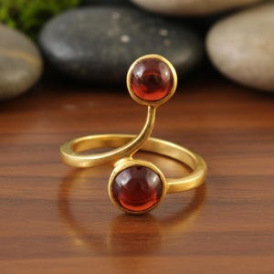Op de afbeelding: Een goudkleurige ring met twee ronde, rode edelstenen. De ring heeft een uniek, wervelend ontwerp, met een edelsteen aan de basis en een andere aan de top van een gebogen steel. De edelstenen zijn gezet in een gouden bezel.