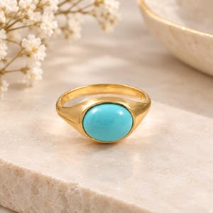 Könnte beinhalten: Ein goldener Ring mit einer glatten, polierten Ringschiene und einem zentralen, ovalen, hellblauen Türkisstein in einer Zargenfassung. Der Ring hat ein massives, abgerundetes Design mit einer glänzenden Oberfläche, die das Licht reflektiert.