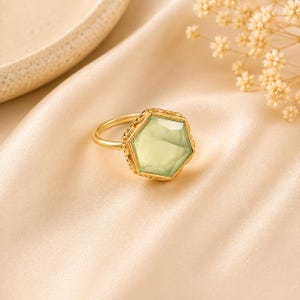 Könnte beinhalten: Ein Goldring mit einem großen, sechseckigen, blassgrünen Edelstein in einer dekorativen Metallfassung. Der Ring ist dünn und kreisförmig, und die Fassung weist filigrane Metallarbeiten entlang der Kanten des sechseckigen Steins auf.