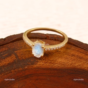 Könnte beinhalten: Ein goldener Ring mit einem ovalen, schillernden Mondstein-Mittelstein. Das Band ist goldfarben mit kleinen, klaren Akzentsteinen, die oben eingesetzt sind. Der Ring wird auf einer Holzoberfläche präsentiert.