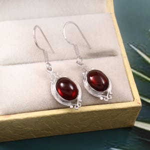 May include: A pair of silver earrings with oval, deep red gemstones. The earrings have a decorative, openwork design around the gemstones and a simple hook closure. The earrings are displayed in a gold-colored jewelry box.