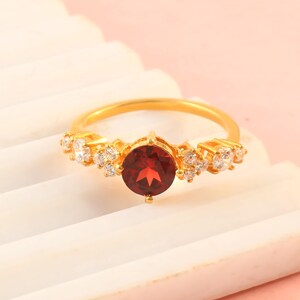 Könnte beinhalten: Ein Goldring mit einem zentralen, rund geschliffenen, tiefroten Granat-Edelstein. Die Ringschiene ist auf beiden Seiten mit Clustern aus kleinen, klaren, rund geschliffenen weißen Steinen besetzt, was ein symmetrisches Design mit Goldfinish ergibt.