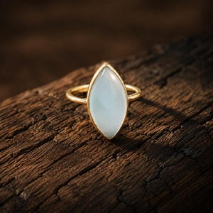 Könnte beinhalten: Ein goldener Ring mit einem glatten, durchscheinenden weißen Stein im Marquise-Schliff, der in einer einfachen Metallschiene gefasst ist. Der Stein hat ein weiches, milchiges Aussehen und wird von einer dünnen Goldfassung gehalten.