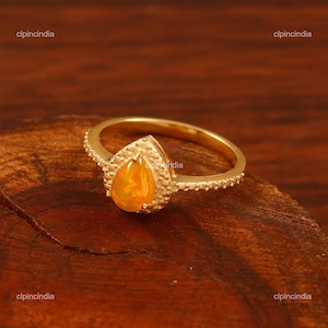 Könnte beinhalten: Ein goldener Ring mit einem birnenförmigen, orangefarbenen Opal in einer texturierten Fassung. Das Ringband ist dünn und poliert, während der Edelstein von einem dekorativen, granulierten Metallrand umgeben ist, der das Design ergänzt.