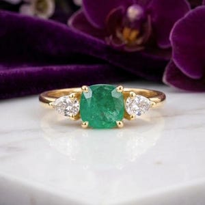 Könnte beinhalten: Ein Goldring mit einem zentralen, kissenförmigen grünen Smaragd, der von zwei tropfenförmigen weißen Diamanten flankiert wird. Der Ring ist auf einem polierten Gelbgoldband gefasst und zeigt ein klassisches Drei-Stein-Design mit Krappenfassungen.