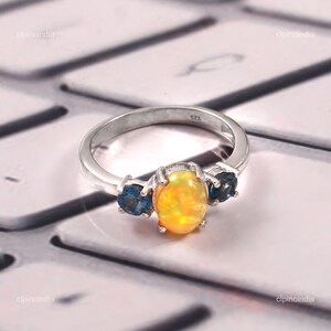 Könnte beinhalten: Ein silberner Ring mit einem ovalen, gelb-orangen Opal und zwei blauen Edelsteinen. Der Ring liegt auf einer hellen Oberfläche. Das Design des Rings ist elegant.