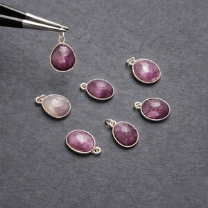 May include: A collection of gemstone pendants with a silver-colored setting. The pendants feature a faceted, oval-shaped, and teardrop-shaped, deep pink gemstone. One pendant is held by tweezers, while the others are scattered on a dark gray surface.