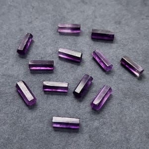 May include: A collection of eleven small, rectangular, translucent purple gemstone beads. Each bead features a polished, elongated shape with flat surfaces and sharp edges, displaying a deep violet hue that varies in intensity across the pieces.