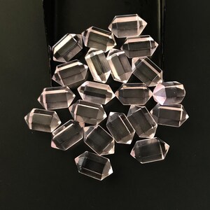 Puede incluir: Una colección de pequeños cristales de cuarzo rosa de doble terminación con un aspecto translúcido de color rosa pálido. Cada cristal presenta una forma hexagonal facetada con extremos puntiagudos, dispuestos en un grupo sobre un fondo oscuro.