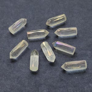 May include: Eight small, clear, faceted crystal points arranged in a circular pattern. Each piece features a pointed tip and a cylindrical body with a smooth, reflective surface that displays subtle iridescent highlights across the transparent material.