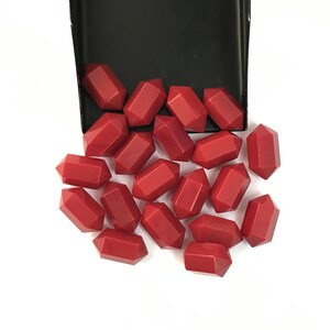 May include: A collection of small, solid red beads with a double-pointed, hexagonal shape. These beads are scattered below a black rectangular container, displaying a smooth, matte finish and uniform geometric design across all individual pieces.