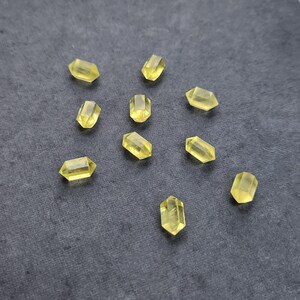 May include: A scattering of small, faceted, yellow crystal beads on a dark grey surface. The crystals are rectangular with pointed ends and appear translucent. Each bead is approximately 1 cm in length.