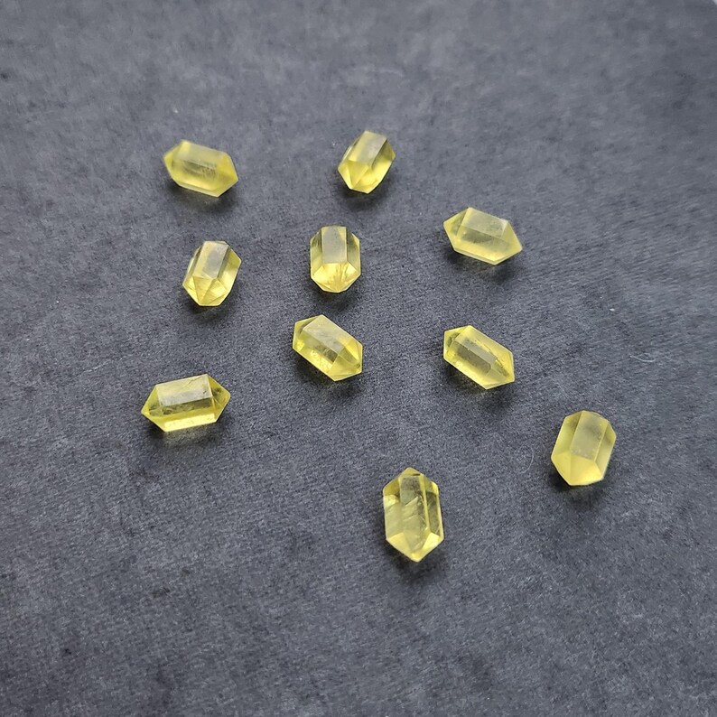 May include: A scattering of small, faceted, yellow crystal beads on a dark grey surface. The crystals are rectangular with pointed ends and appear translucent. Each bead is approximately 1 cm in length.