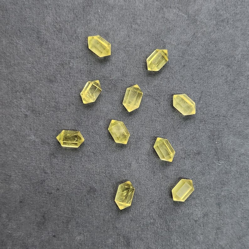 May include: Ten yellow crystal gemstones, each with a hexagonal shape and pointed ends, are scattered on a dark grey surface. The crystals' translucent quality is evident. These gemstones are suitable for jewellery making or collecting, showcasing their natural beauty.