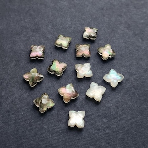 May include: A collection of twelve small, four-petaled flower-shaped beads made of iridescent mother-of-pearl. Each bead features a faceted surface that reflects light with subtle pink, green, and white tones, arranged in a scattered pattern.