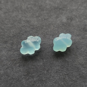May include: Two small, translucent, light blue beads shaped like stylised clouds. Each bead features a smooth, rounded surface with a glossy finish that reflects light, creating a soft, pale blue appearance with subtle variations in colour depth.