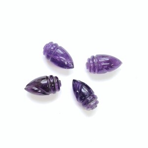 May include: Four carved amethyst gemstone bullet-shaped beads. Each bead has a pointed end and a ridged, cylindrical base. The gemstones are a deep purple color and are displayed against a white background.