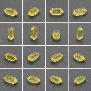 May include: A grid of yellow crystal beads. Each bead is an elongated hexagonal prism with pointed ends, displaying a translucent quality. The crystals have a faceted appearance, reflecting light. The background is a dark grey surface.