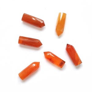 Op de afbeelding: Zes gefacetteerde carneool edelstenen in de vorm van kleine, gepolijste punten. Elke steen heeft een doorschijnende, levendige oranje-rode tint met gladde, geometrische oppervlakken die aan één kant toelopen in een punt, verspreid gerangschikt.