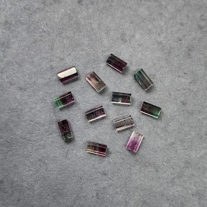 Puede incluir: Una colección de cristales de turmalina rectangulares, que muestran una mezcla de colores verdes, morados y transparentes. Los cristales están dispersos sobre una superficie gris, mostrando su apariencia natural y pulida. Estas piedras preciosas son ideales para la fabricación de joyas.