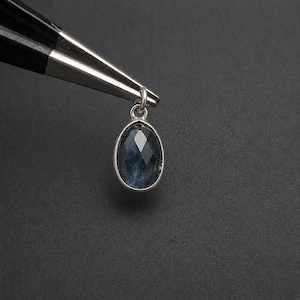 Puede incluir: Colgante de piedra preciosa azul ovalada y facetada con engaste de plata. El colgante está suspendido por un lazo plateado y sujeto por unas pinzas plateadas sobre un fondo oscuro. La piedra preciosa es de color azul intenso.