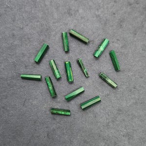 Può includere: Una collezione di piccole perline di malachite verde. Le perline sono di forma esagonale e mostrano una varietà di tonalità e motivi verdi. Le perline sono sparse su una superficie grigio scuro, creando una disposizione visivamente accattivante.