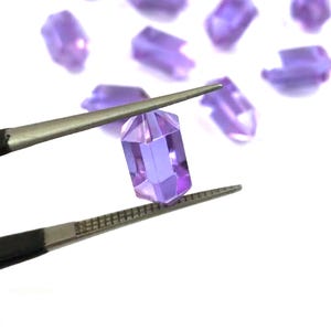 May include: A small, translucent, light purple, rectangular-cut gemstone held by metal tweezers. The gem features faceted edges that catch the light, creating a clear, polished appearance. Several identical loose purple gems are scattered nearby.