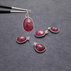 May include: Four oval-shaped, faceted deep red ruby pendants set in silver bezels with small jump rings. One pendant is held by black metal tweezers, while the other three are arranged below on a dark gray textured surface.