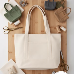 Könnte beinhalten: Ein cremefarbenes Schnittmuster für eine Tragetasche mit dem Text "HANDMADE TOTE BAG PATTERN" und "Craft Your Perfect Everyday Bag". Das Bild enthält Miniatur-Tragetaschen in verschiedenen Farben, Scheren und Nähzubehör. Der Text "EASY Step-bn-Step Instructions Included!" ist ebenfalls sichtbar.