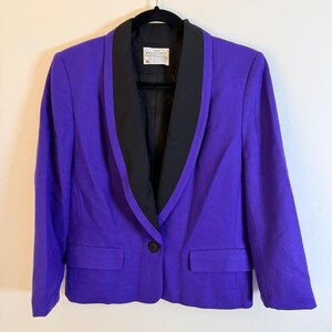 Pode incluir: Um blazer roxo vibrante com lapela preta e um único botão preto. O blazer apresenta dois bolsos frontais e um forro interno preto. A peça de roupa está pendurada em um cabide preto.
