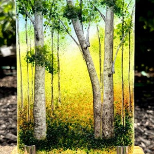 May include: A rectangular fused glass panel depicting a forest scene with white birch trees, green foliage, and a bright yellow background. The glass art piece is held upright by two silver metal clips attached to a rectangular wooden base.