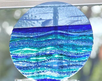 May include: A circular fused glass art piece featuring a horizontal composition of layered colors. The top section is a solid light blue, while the lower portion displays wavy, textured bands of deep navy, royal blue, and bright turquoise glass.