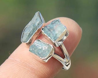 May include: A silver ring featuring three raw, light blue aquamarine stones. The stones are set in individual square and rectangular bezels, creating a unique and naturalistic design. The ring is shown on a finger against a blurred green background.