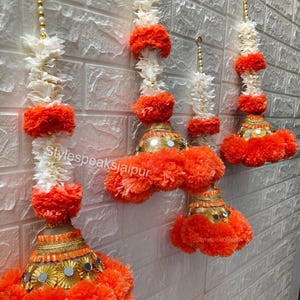 May include: Decorative hanging ornaments with white and orange pom-poms, gold accents, and mirrored details. The ornaments are suspended against a white brick wall. The text "Stylespeaksjaipur" is visible on the ornaments.