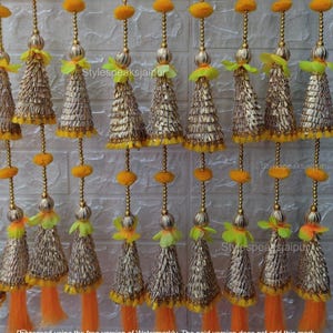 May include: Decorative gold and yellow hanging ornaments. Each ornament features a cone-shaped tassel with gold accents, orange and yellow accents, and a string of gold beads. The ornaments are arranged in rows against a white brick wall.